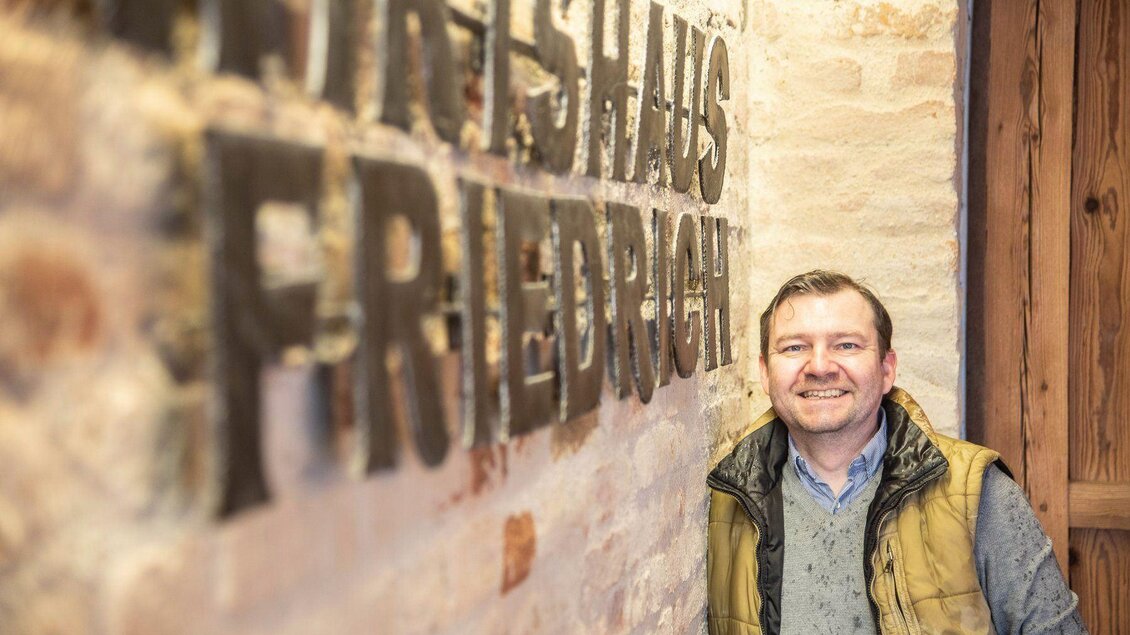 Ein Mann lächelt neben einer Wand mit dem Schriftzug „Friedrichs Haus“. Die Wand hat einen rustikalen Steinlook und vermittelt eine gemütliche Atmosphäre. | © Friedrich