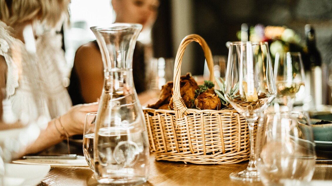Ein gedeckter Tisch mit einem Korb voller Essen und Gläsern. Im Hintergrund sind Menschen zu sehen, die gemeinsam essen und sich unterhalten. | © Wirtshaus Thorbauer - Julian Wallner