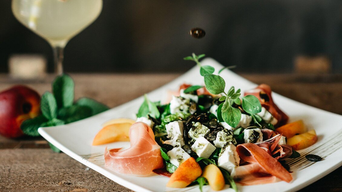 Ein frischer Salat mit Schinken, Feta und fruchtigen Beilagen auf einem weißen Teller. Im Hintergrund ist ein Getränk und eine Apfel zu sehen. | © Wirtshaus Thorbauer - Julian Wallner