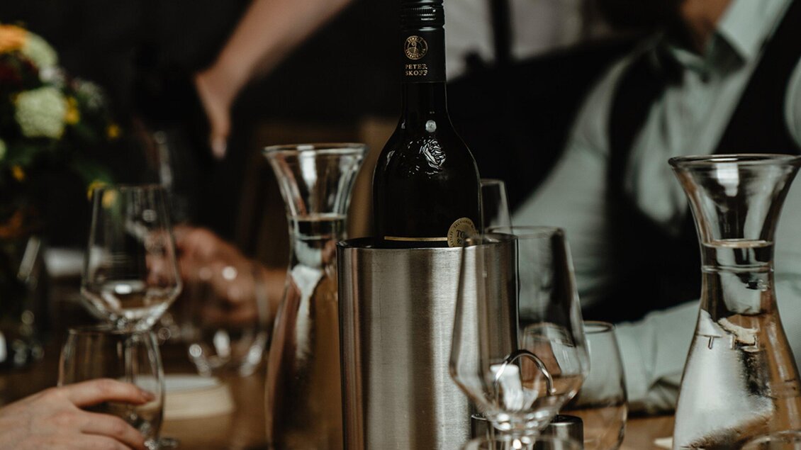 Ein Tisch gedeckt mit Gläsern und Karaffen, auf dem ein Weinflasche in einem Kühler steht. Im Hintergrund sind Hände und Personen sichtbar, die an einem geselligen Treffen teilnehmen. | © Wirtshaus Thorbauer - Julian Wallner