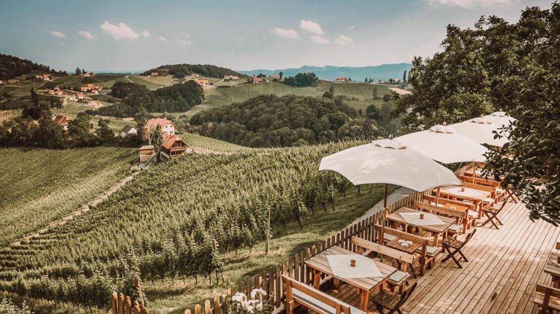 Eine malerische Terrasse mit Tischen und Schirmen, umgeben von sanften Hügeln und Weinbergen. Im Hintergrund sind kleine Häuser und eine grüne Landschaft zu sehen.