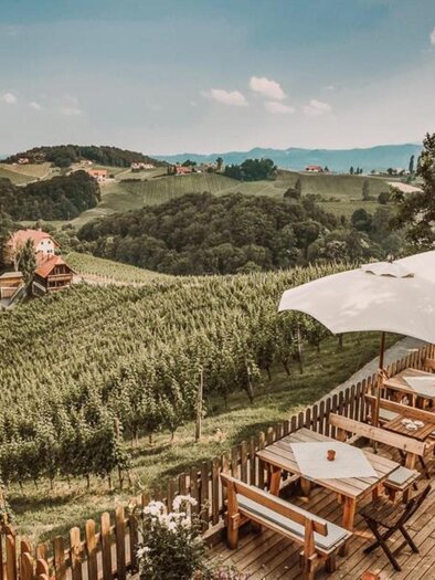 Eine malerische Terrasse mit Tischen und Schirmen, umgeben von sanften Hügeln und Weinbergen. Im Hintergrund sind kleine Häuser und eine grüne Landschaft zu sehen.