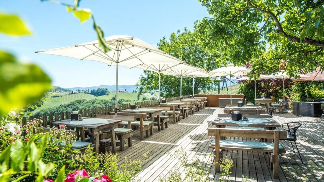 Ein einladender Außenbereich eines Restaurants mit Holzstühlen und Tischen. Sonnenschirme bieten Schatten und die Umgebung ist grün und gepflegt. | © Magdalena Koinegg