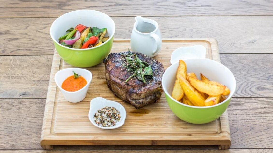 Ein köstliches Gericht mit einem Stück Steak, serviert mit Kartoffelspalten und einem bunten Salat. Dazu gibt es verschiedene Saucen in kleinen Schalen. | © Magdalena Koinegg