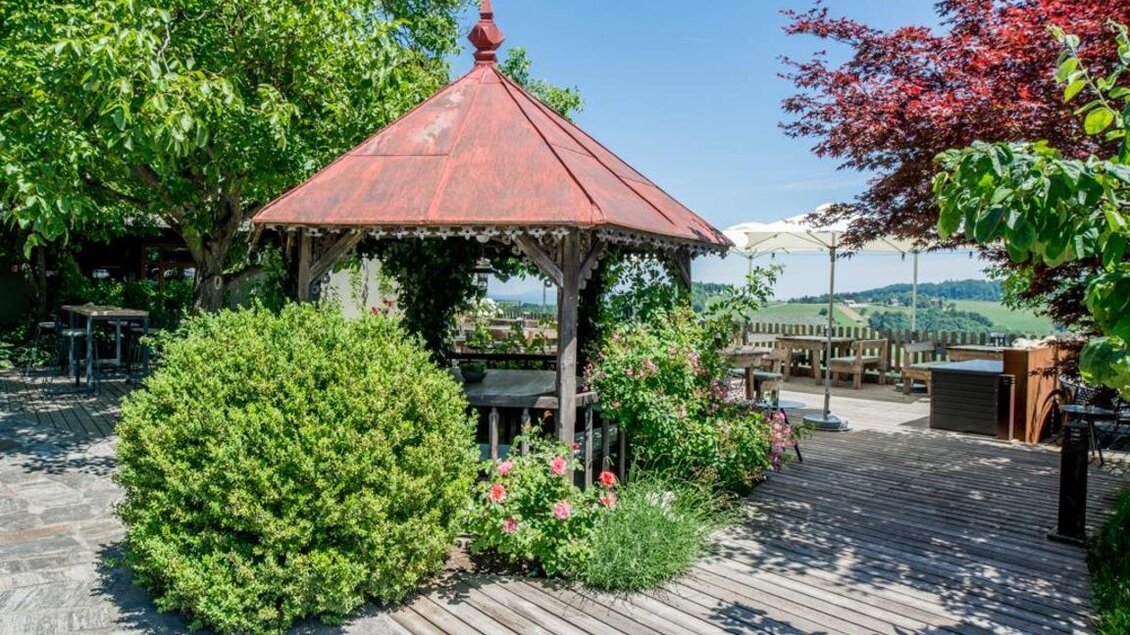 Ein schöner Pavillon in einem Garten mit üppigem Grün und bunten Blumen. Im Hintergrund sind sonnige Plätze zum Entspannen und eine malerische Landschaft zu sehen. | © Magdalena Koinegg