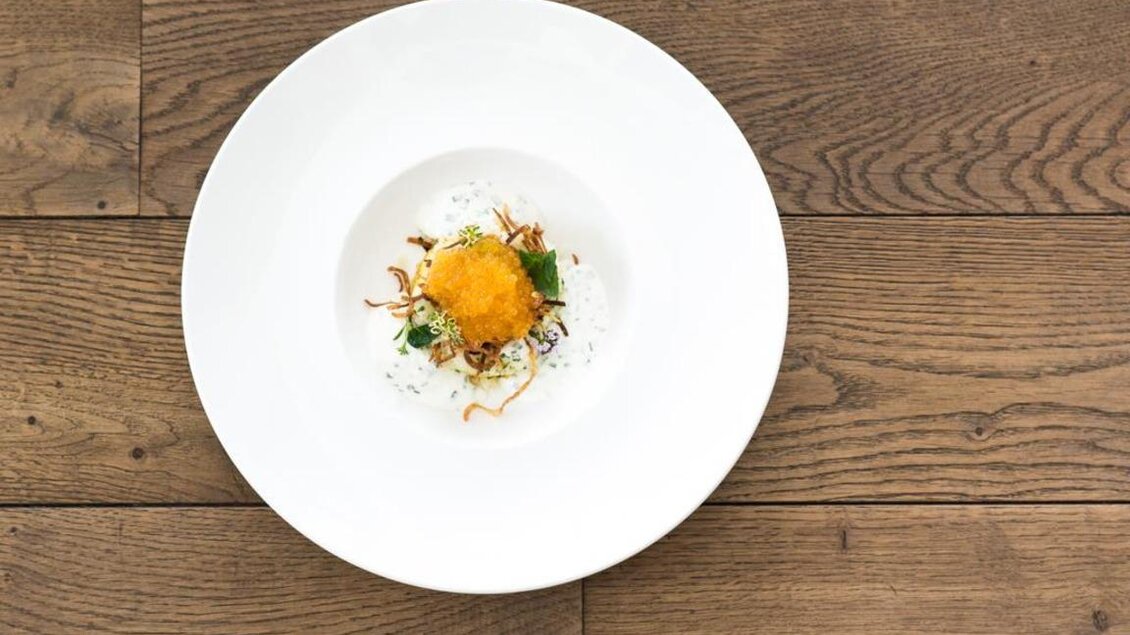 Ein elegantes Gericht auf einem weißen Teller, bestehend aus einer goldenen Kugel, frischen Kräutern und einer dekorativen Beigabe. Der Hintergrund zeigt einen warmen Holztisch. | © Magdalena Koinegg