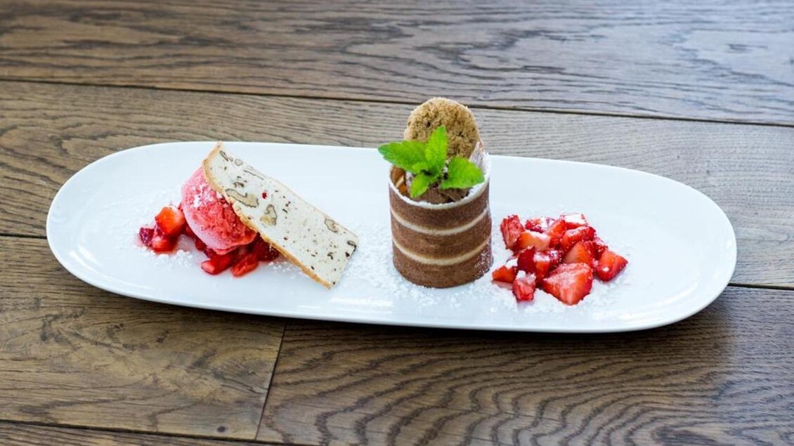 Ein elegantes Dessert auf einem weißen Teller, bestehend aus einer Schokoladenrolle, einer Biskuit-Schnitte und frischen Erdbeeren. Das Gericht ist mit einem Blatt Minze garniert und auf einem Holzuntergrund angerichtet. | © Magdalena Koinegg