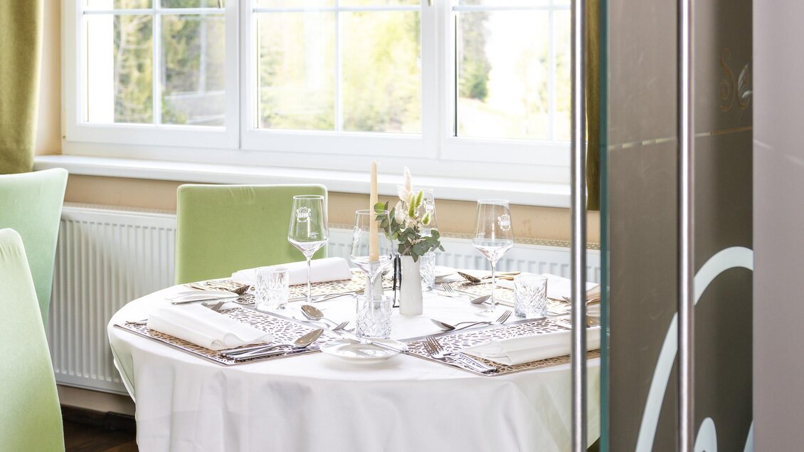 Ein eleganter Tisch mit weißer Tischdecke und edlem Service in einem gemütlichen Raum. Große Fenster lassen viel Tageslicht herein.