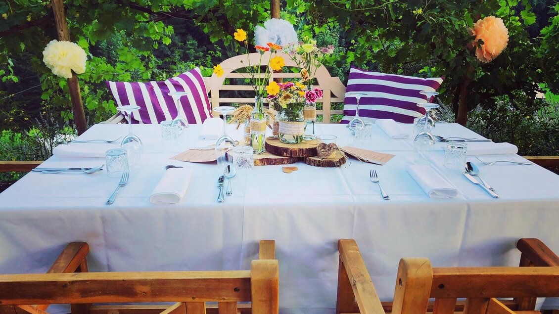 Ein hübsch eingedeckter Tisch unter Weinreben mit bunten Blumen und Kissen. Die Holzstühle laden zum Verweilen ein. | © Landhotel am Gut Moser | Familie Kapun
