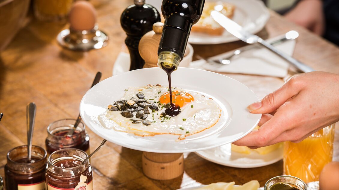 Ein Teller mit einem Spiegelei, das mit Olivenöl beträufelt wird. Im Hintergrund sind verschiedene Frühstückszutaten und Marmeladengläser zu sehen.