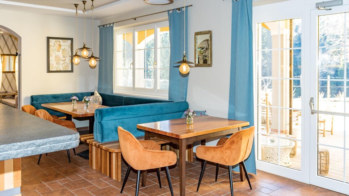 Ein modernes Café mit gemütlichen Sitzplätzen, blauen Vorhängen und warmem Licht. Große Fenster sorgen für einen hellen, einladenden Raum.