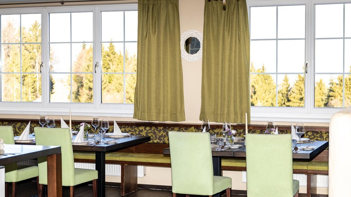 Ein modernes Restaurant mit eleganten Tischen und grünen Stühlen. Große Fenster sorgen für viel Tageslicht und einen Blick auf die Natur.