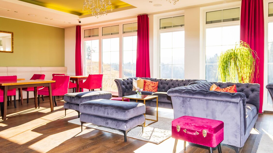 Ein stilvoll eingerichteter Raum mit bequemen, blauen Sofas und roten Vorhängen. Große Fenster sorgen für viel Tageslicht und bieten einen schönen Ausblick.