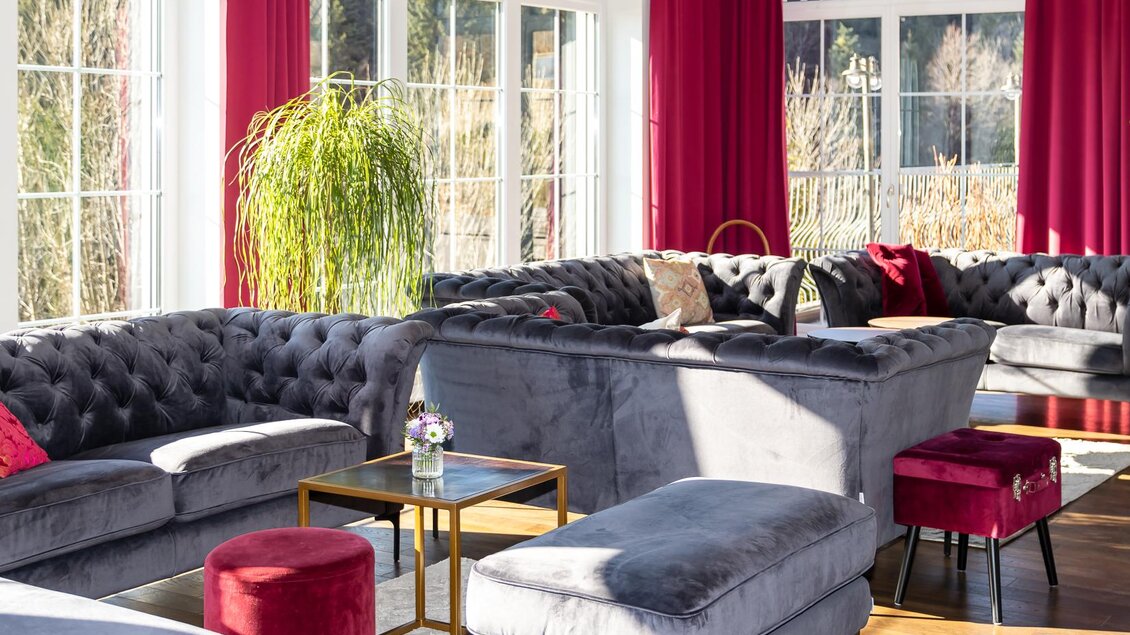 Ein elegantes Wohnzimmer mit grauen Samtsofas und roten Akzenten. Große Fenster lassen viel Licht herein und bieten einen Blick auf die Natur.