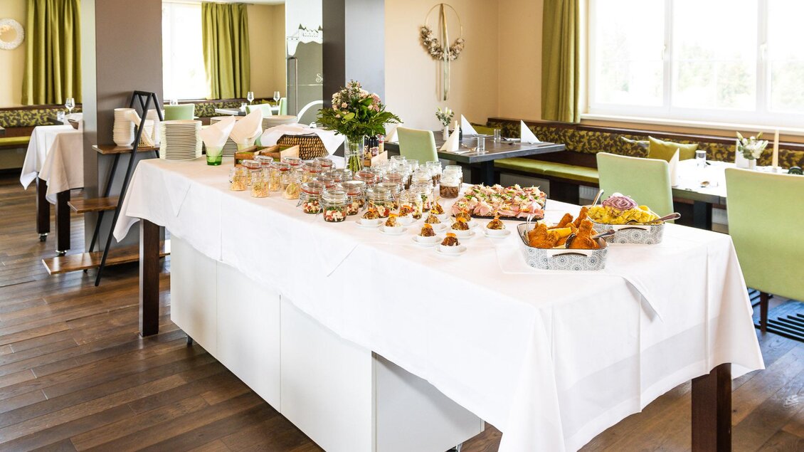 Ein elegantes Restaurant mit einem gedeckten Tisch, der mit verschiedenen Speisen und Getränken dekoriert ist. Die Einrichtung ist hell und freundlich mit grünen Vorhängen und Holzfußboden.