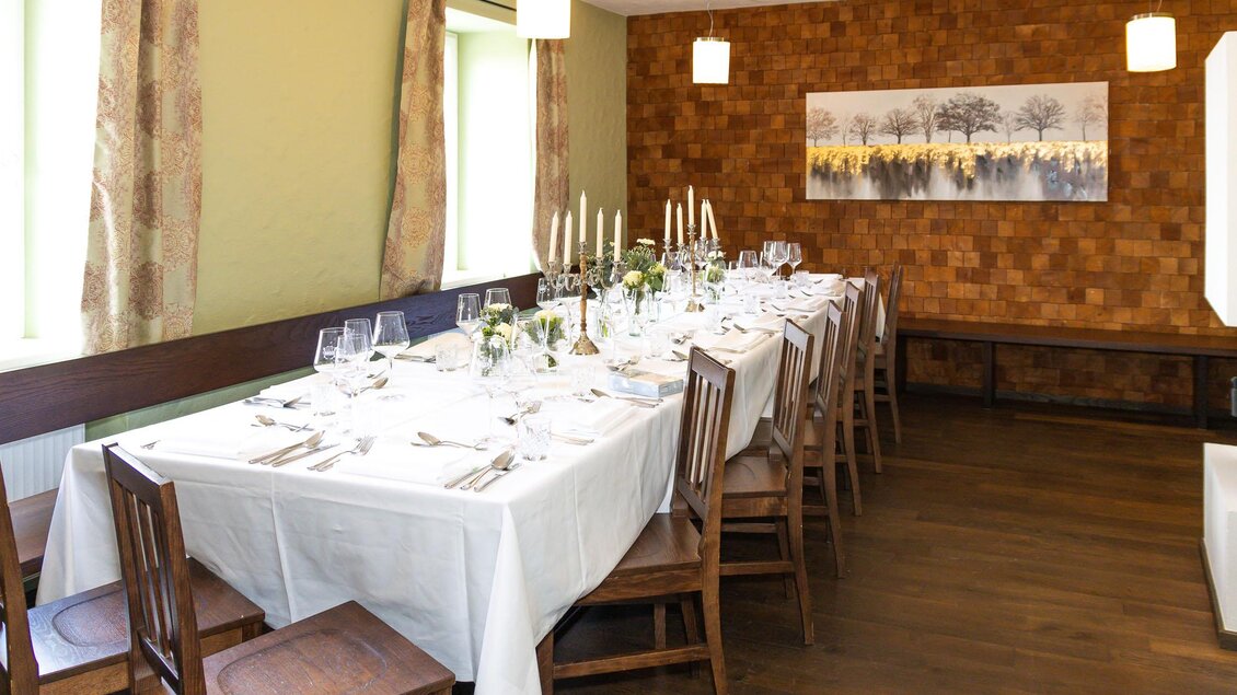 Ein elegantes Esszimmer mit einem lange Tisch, der für ein Festmahl gedeckt ist. Große Fenster lassen viel Licht herein und schaffen eine gemütliche Atmosphäre.
