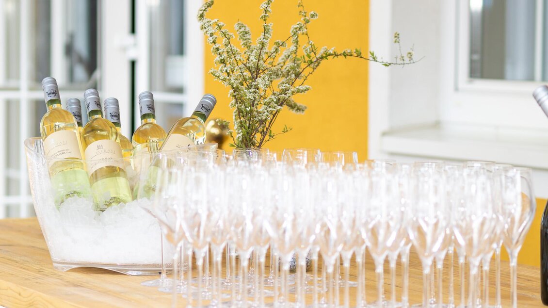 Eine elegante Präsentation von Weingläsern und Flaschen Wein, die auf einem Holztablett angeordnet sind. Im Hintergrund ist eine frische Dekoration aus Zweigen zu sehen.