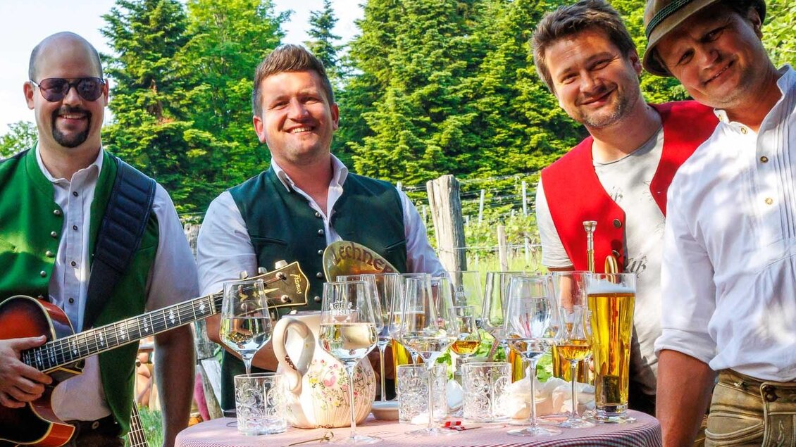 Eine Gruppe von vier Männern in traditioneller Tracht steht um einen Tisch mit Gläsern und Getränken. Im Hintergrund sind Bäume und eine grüne Landschaft zu sehen.