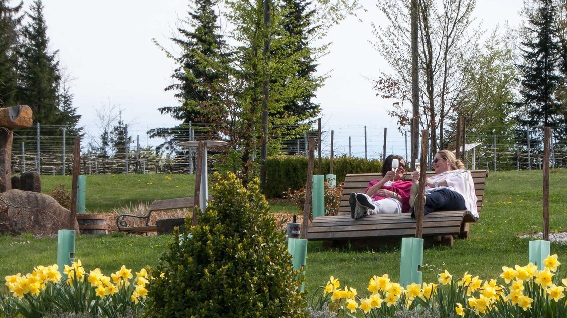 Ein schöner Garten mit gelben Narzissen und einer grünen Wiese. Zwei Personen entspannen auf einer Bank unter Bäumen.