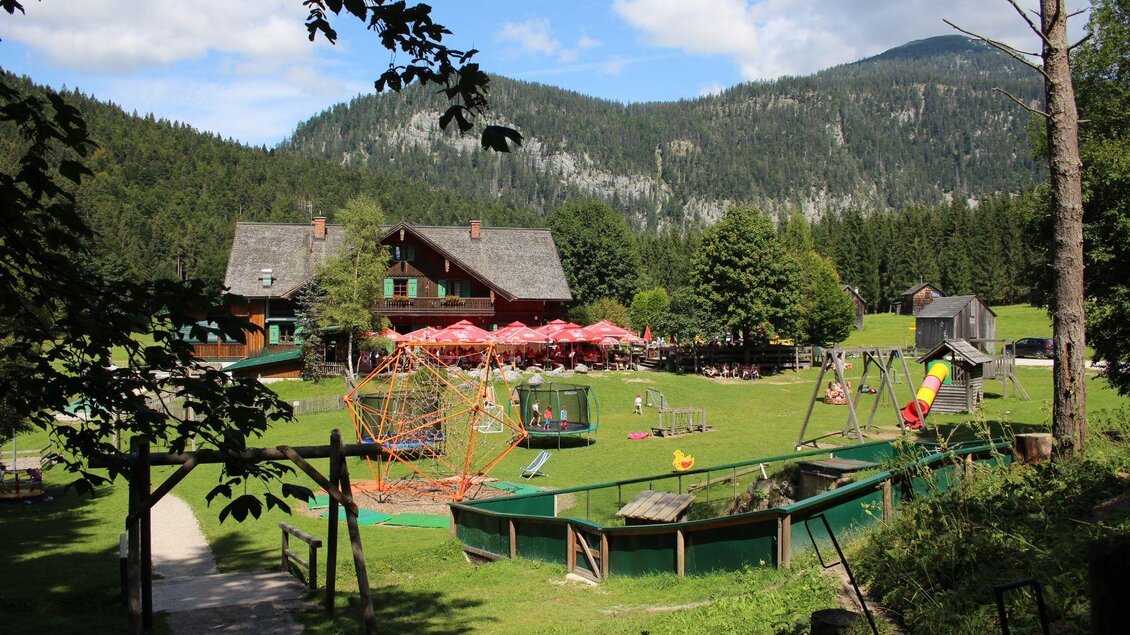 Eine malerische Landschaft mit einem Spielplatz und einem großen Haus im Hintergrund. Die Umgebung ist von Bäumen und Bergen umgeben, ideal für Familienaktivitäten. | © Viola Lechner