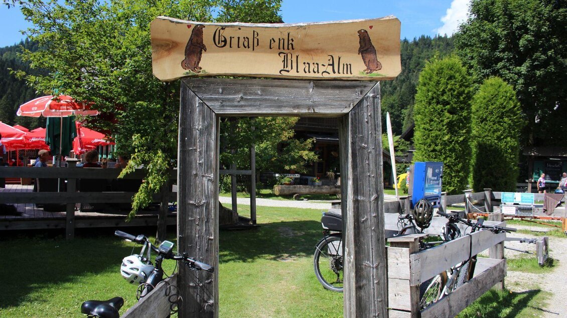 Ein Holzschild mit der Aufschrift „Grüß dich, PrazAlm“ steht am Eingang. Im Hintergrund sind Bäume und eine Terrasse mit roten Sonnenschirmen zu sehen. | © Viola Lechner