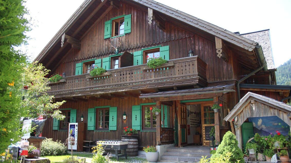 Ein traditionelles Holzhaus mit grünen Fensterläden und einem Balkon. Umgeben von einem gepflegten Garten und Bäumen. | © Viola Lechner