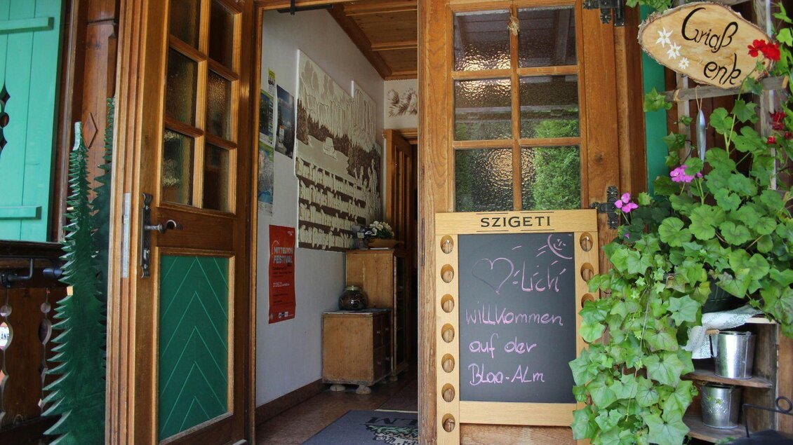 Ein einladender Eingang zu einem gemütlichen Gasthaus mit einer Tafel, auf der „Willkommen auf der Bloa-Alm“ steht. Die Tür ist offen und es wachsen Blumen auf der Seite. | © Viola Lechner