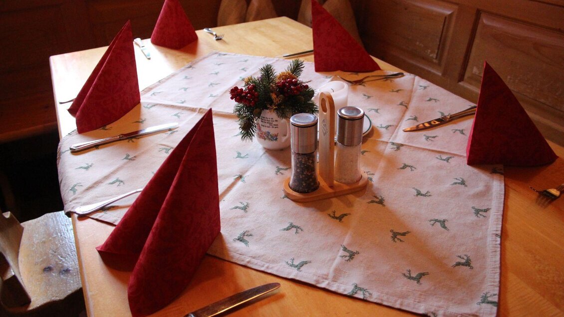 Ein festlich gedeckter Tisch mit roten Servietten und einem dekorativen Mittelpunkt. Die Tischdecke zeigt ein schlichtes Muster und das Besteck ist ordentlich angerichtet. | © Viola Lechner