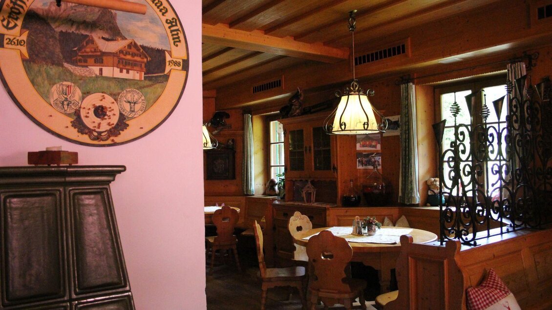 Ein gemütlicher Innenraum mit rustikalen Holzstühlen und einem traditionellen Holzofen. An der Wand hängt ein dekorativer Teller, der eine ländliche Szene darstellt. | © Viola Lechner