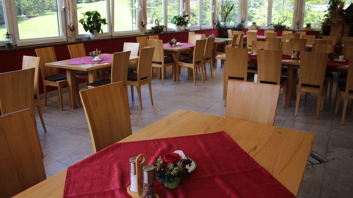 Ein heller Restaurantbereich mit Holzstühlen und Tischen. Die Tische sind mit roten Tischdecken dekoriert und bieten einen Blick auf die grüne Landschaft. | © Viola Lechner