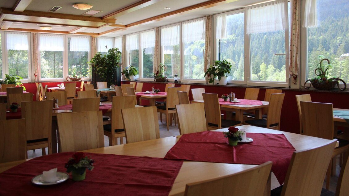 Ein gemütlicher Restaurantbereich mit Holzstühlen und Tischen, die mit roten Tischdecken gedeckt sind. Große Fenster bieten einen Blick auf die grüne Landschaft draußen. | © Viola Lechner