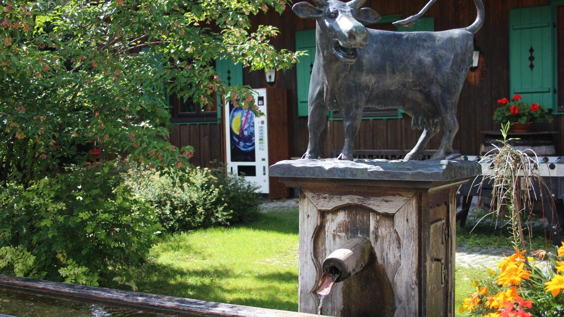 Eine Bronze-Statue einer Kuh steht auf einem Brunnen in einer grünen Umgebung. Im Hintergrund sind bunte Blumen und ein Holzhaus zu sehen. | © Viola Lechner