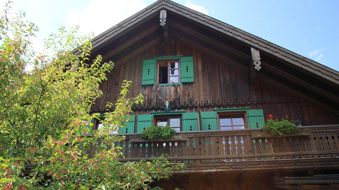 Ein traditionelles Holzhaus mit grünen Fensterläden und einem Balkon. Im Vordergrund sind Bäume und Pflanzen zu sehen. | © Viola Lechner