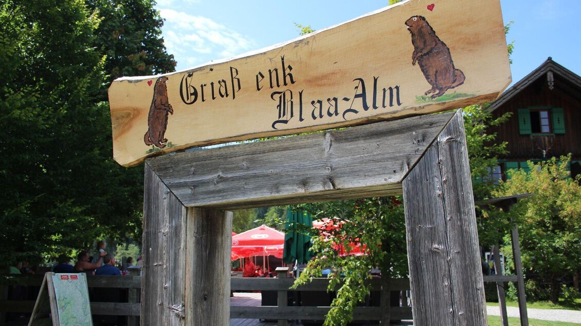 Ein rustikales Schild mit der Aufschrift "Griaß enk Blaa-Alm" und einem Bärenmotiv. Die Umgebung ist grün und einladend, mit mehreren roten Zelten im Hintergrund. | © Viola Lechner