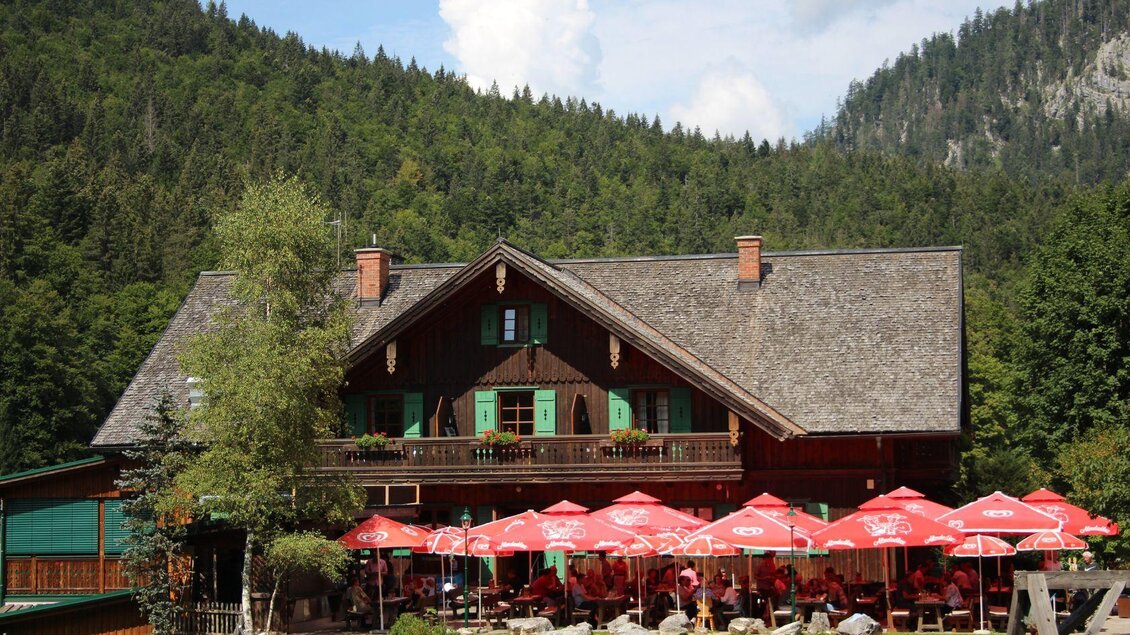 Ein traditionelles Holzhaus mit einem großen Garten und Terrassen. Rote Sonnenschirme laden zum Verweilen ein, umgeben von einer grünen Waldlandschaft. | © Viola Lechner