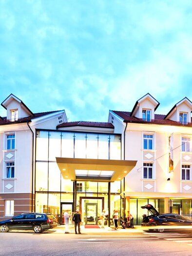 A modern hotel building with an inviting facade. Several vehicles are parked in front of the hotel, and the sky has a soft blue hue. | © Stainzerhof KG
