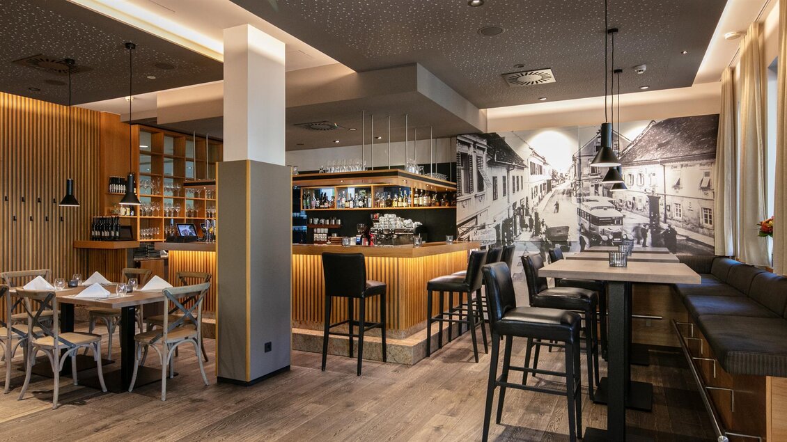 Ein modernes Restaurant mit einer stilvollen Bar und eleganten Sitzgelegenheiten. Die Wände sind mit Holz und einem historischen Bild dekoriert. | © Stainzerhof KG