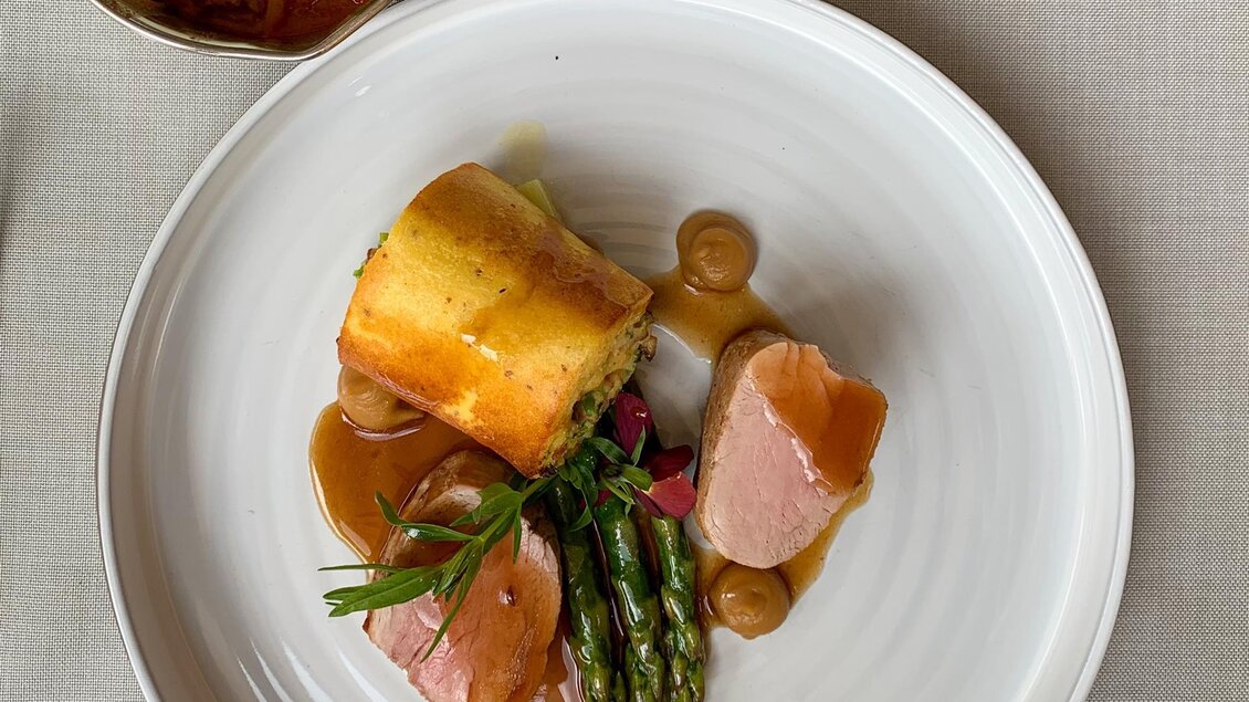 Ein eleganter Teller mit zartem Fleisch, grünem Spargel und einer butterartigen Beilage. Im Hintergrund befindet sich eine kleine Schüssel mit Sauce. | © Stainzerhof KG