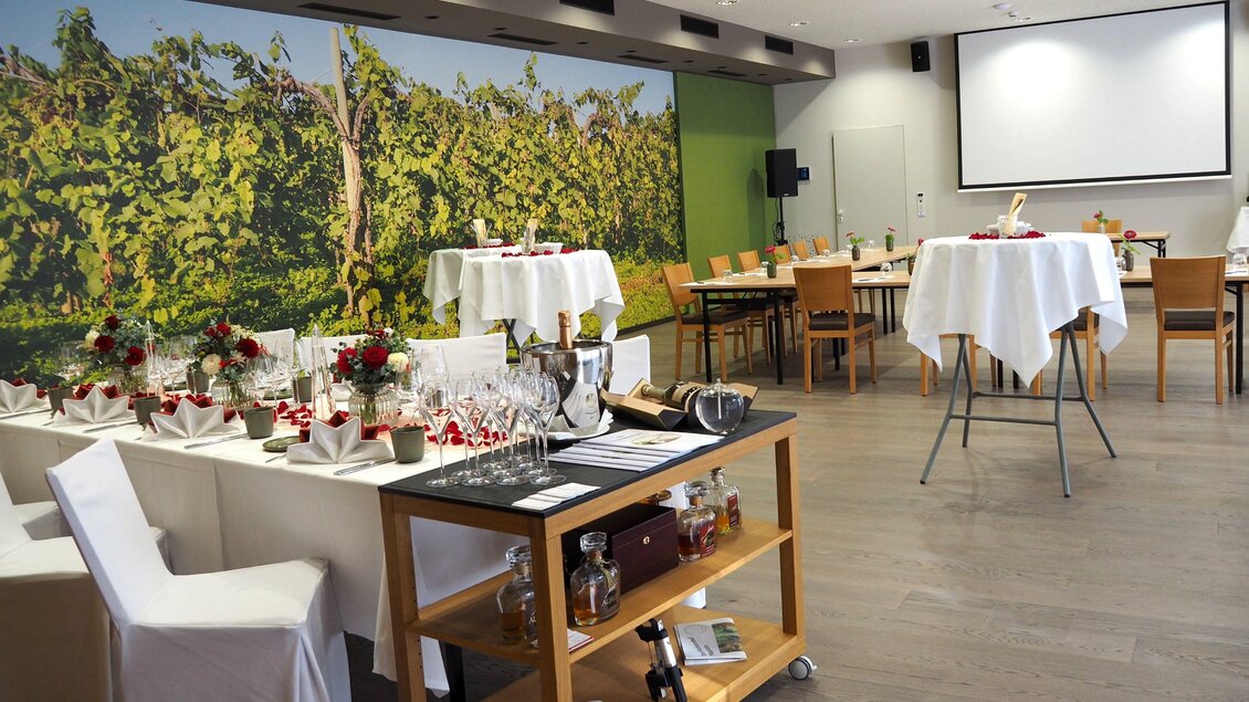 Ein eleganter Veranstaltungsraum mit Tischen, gedeckt für ein Festmahl. Im Hintergrund ist eine Wand mit einem Bild von Weinreben zu sehen. | © Renate Pein