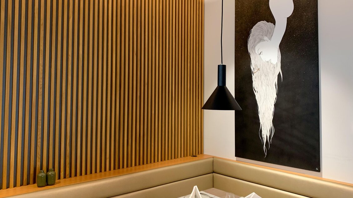 Ein eleganter Restaurantbereich mit Holzvertäfelung und modernem Kunstwerk an der Wand. Die Tischdeko besteht aus weißen Servietten und schwarzen Kerzenhaltern. | © Renate Pein