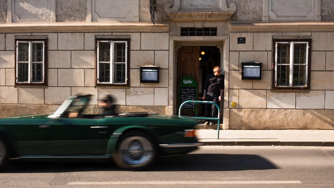 Ein Mann steht in der Tür eines charmanten Gebäudes, während ein grünes Auto vorbeifährt. Die Architektur des Hauses ist klassisch und gut erhalten. | © Robert Bodnar