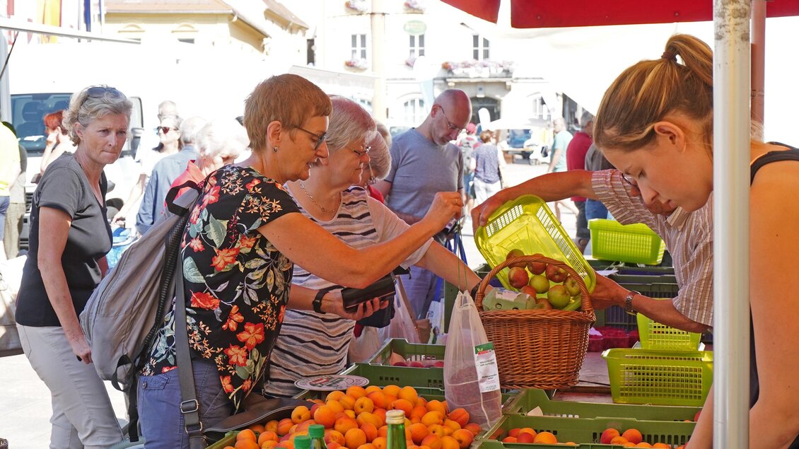 Ein lebhafter Markt mit Menschen, die frisches Obst einkaufen. Die Stände sind reichhaltig mit Orangen und anderen Produkten gefüllt. | © Stadtgemeinde Bruck/Foto: Maili