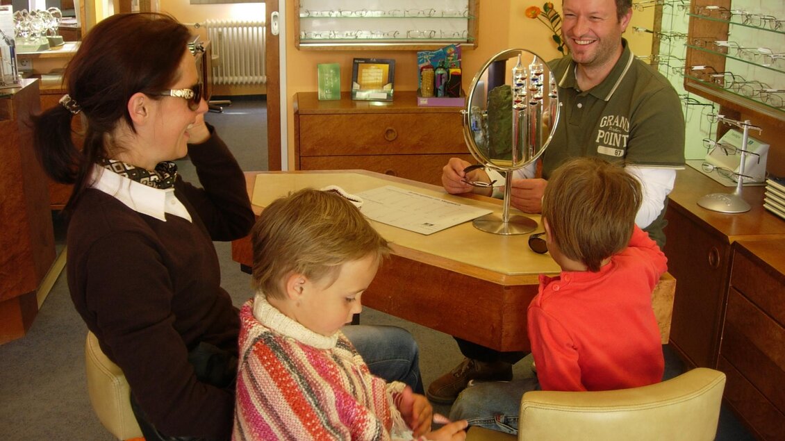 Ein Mann berät eine Familie in einem Optikergeschäft. Die Mutter sitzt mit einer Sonnenbrille, während die Kinder aufmerksam zuhören. | © Wutscher Optik