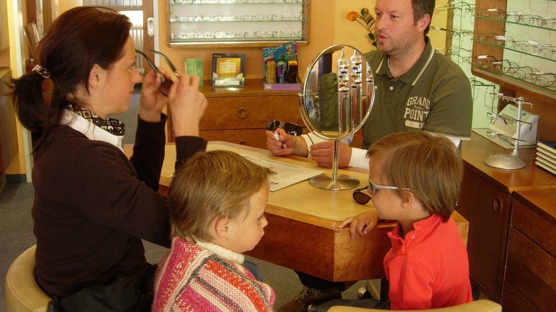 Ein Mann berät eine Frau und zwei Kinder in einem Brillengeschäft. Auf dem Tisch stehen eine Handspiegel und verschiedene Brillen. | © Wutscher Optik