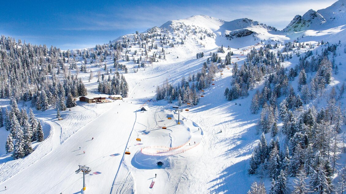 Eine wunderschöne verschneite Berglandschaft mit Skihütten und vielen Bäumen. Skipisten mit Skifahrern und einer klaren blauen Himmel. | © Josh Absenger