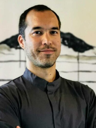A man stands in front of a minimalist mural. He looks directly at the camera and is wearing a dark shirt. | © Yamauchi Ramen