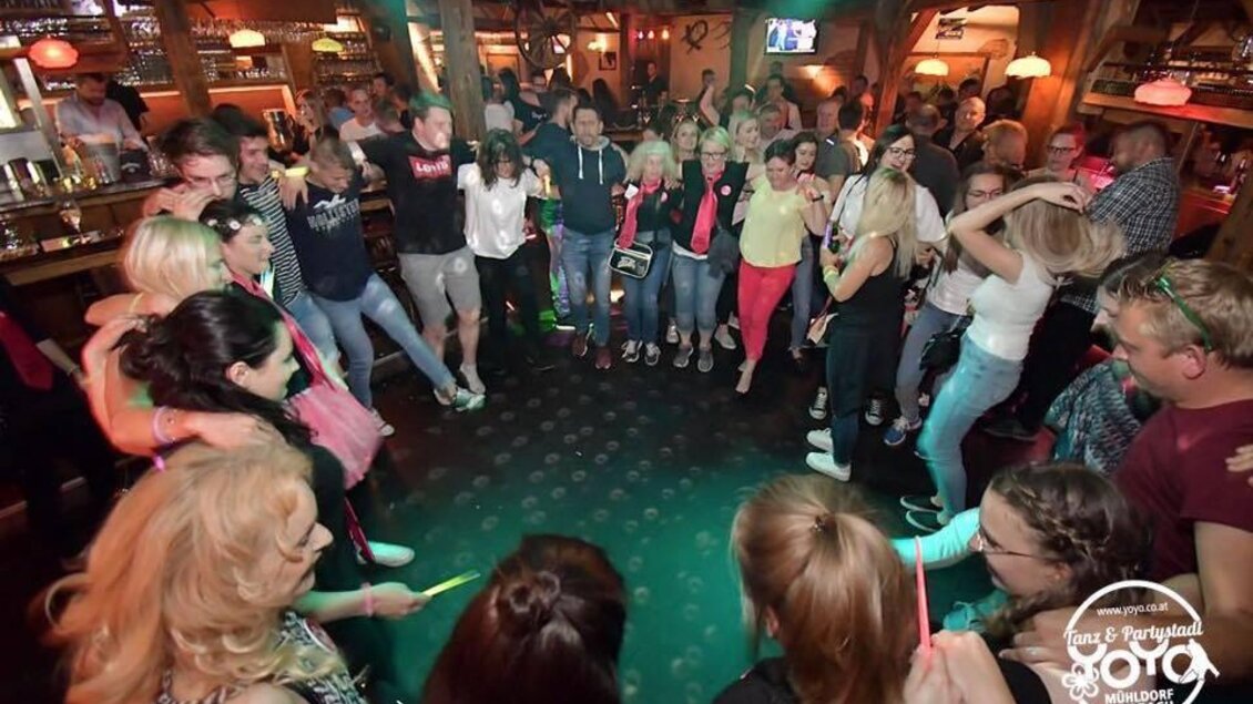 Eine fröhliche Menschenmenge tanzt um einen zentralen Bereich in einer Bar. Die Atmosphäre ist lebhaft und ausgelassen, mit Musik und Lichtern im Hintergrund. | © Yoyo Feldbach