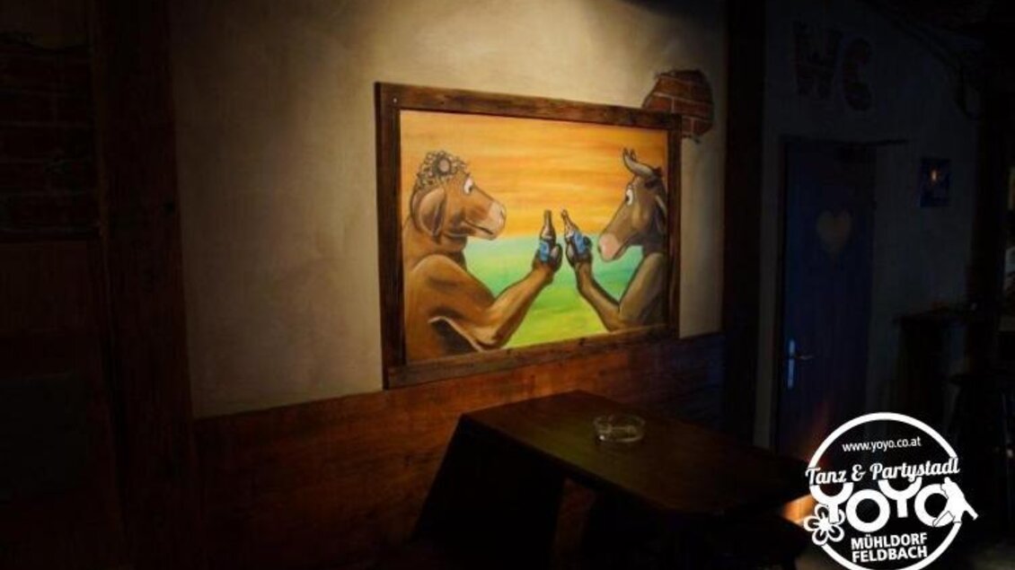 Eine kreative Wandmalerei zeigt zwei Kühe, die mit Bierflaschen anstoßen. Der Raum hat eine rustikale Atmosphäre mit Holzmöbeln. | © Yoyo Feldbach