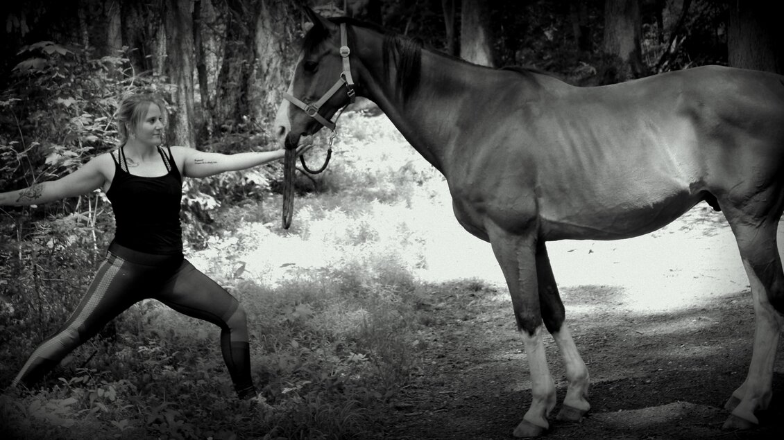 Eine Frau steht in einer Yoga-Position neben einem Pferd im Wald. Die Szene ist in Schwarz-Weiß gehalten und zeigt die Verbindung zwischen Mensch und Tier. | © Michael Größinger