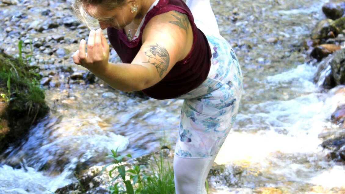 Eine Frau übt eine Yoga-Positur am Ufer eines kleinen Bachs. Der Hintergrund zeigt grünes Laub und fließendes Wasser. | © Michael Größinger
